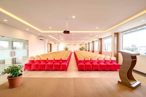 Un salón de conferencias con filas de sillas rojas y color beige, un proyector en el techo y un ventanal grande a la derecha.