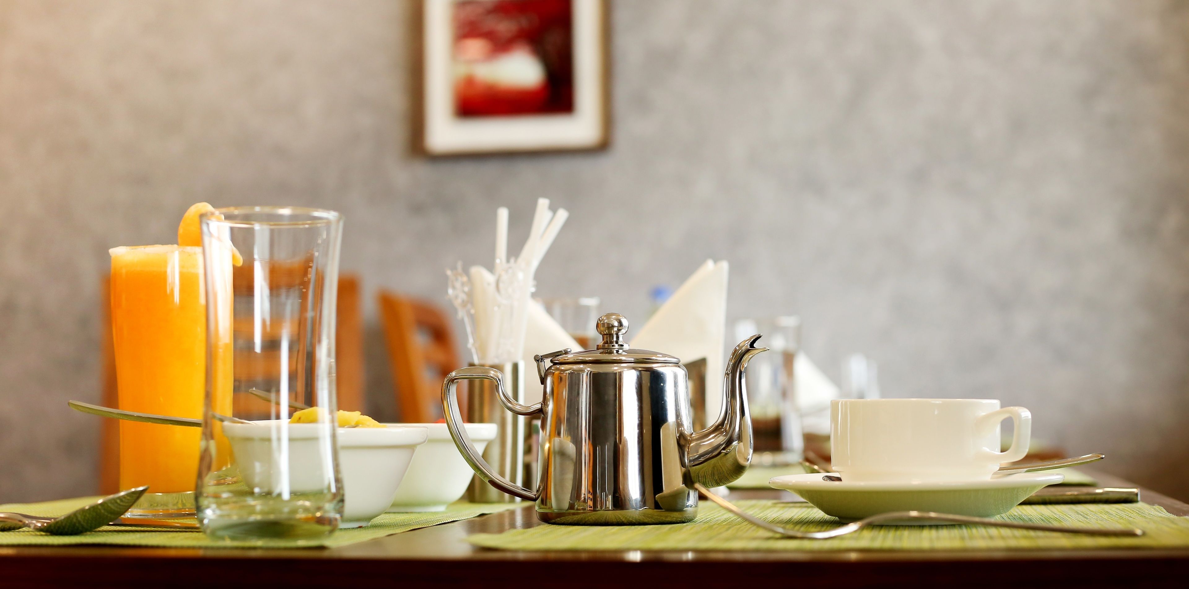 Una mesa de desayuno con una jarrón, tetera, tazas y un vaso de jugo de naranja.