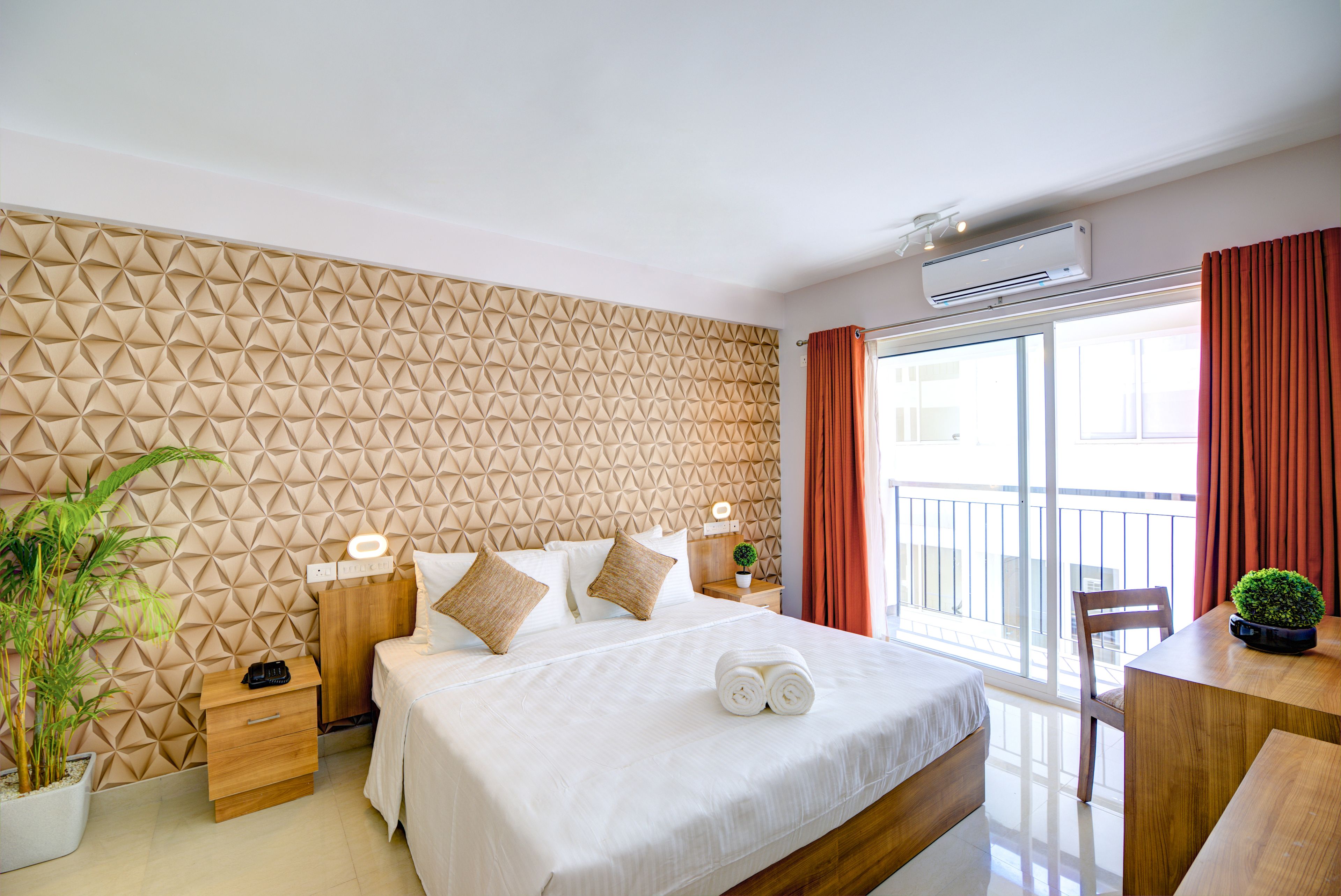 A hotel room with a geometric patterned wall, a bed with white bedding, a bedside table with a lamp, a potted plant, and a view of the outside balcony.