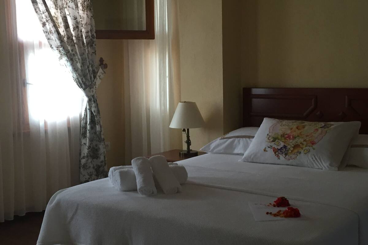 A neatly made bed with white linens and a floral pillow, a bedside lamp, and a window with sheer curtains.
