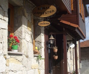 A stone building with a wooden sign reading "KONAK LAPEISTRA" and a phone number.