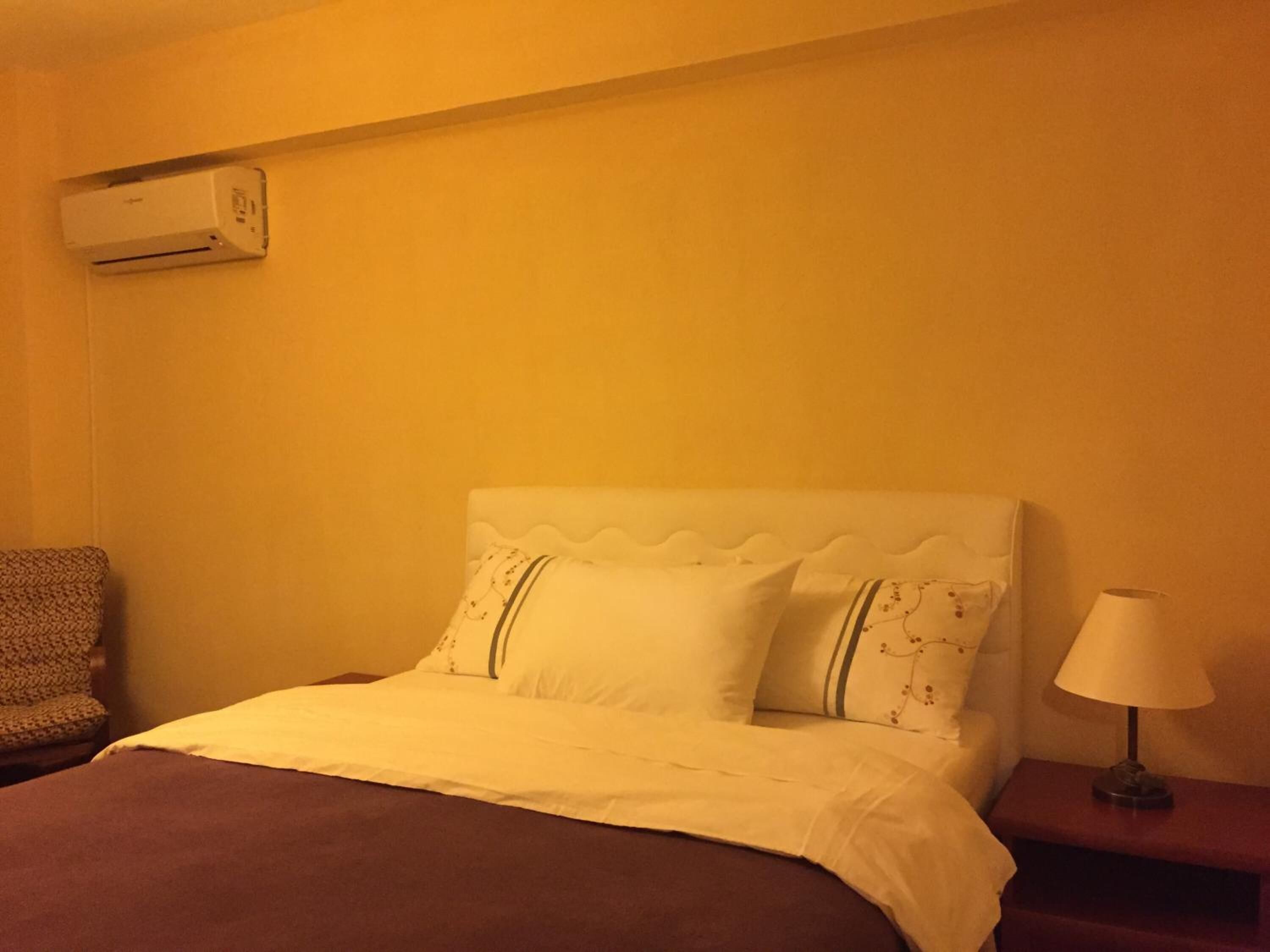 A hotel room with a bed, a chair, a nightstand with a lamp, and an air conditioning unit on the wall.