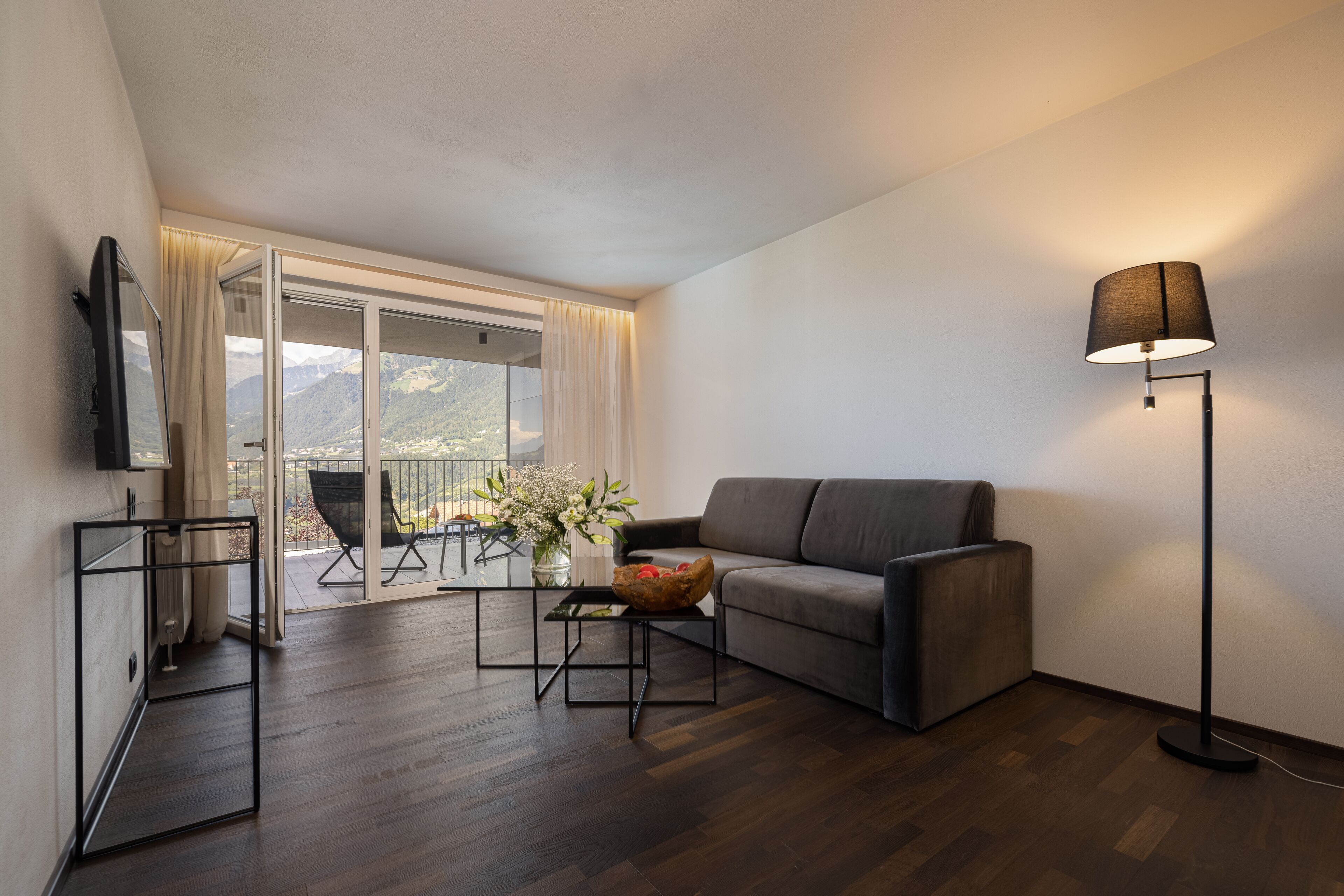 Una sala de estar moderna con un sofá, una mesa de centro con un florero, una lámpara de pie y un televisor sobre un mueble.