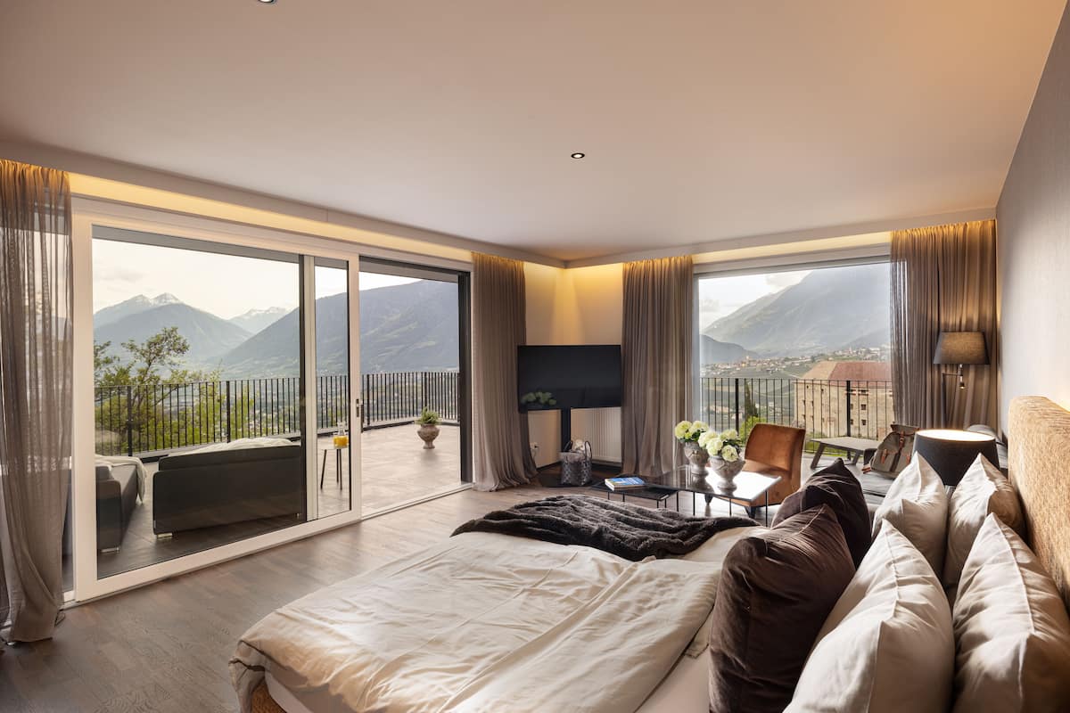 A modern hotel room with a large bed, a sofa, a desk, and a balcony with a mountain view.