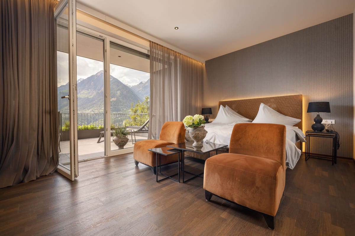 A modern hotel room with a large bed, two armchairs, a small table, and a balcony with a mountain view.