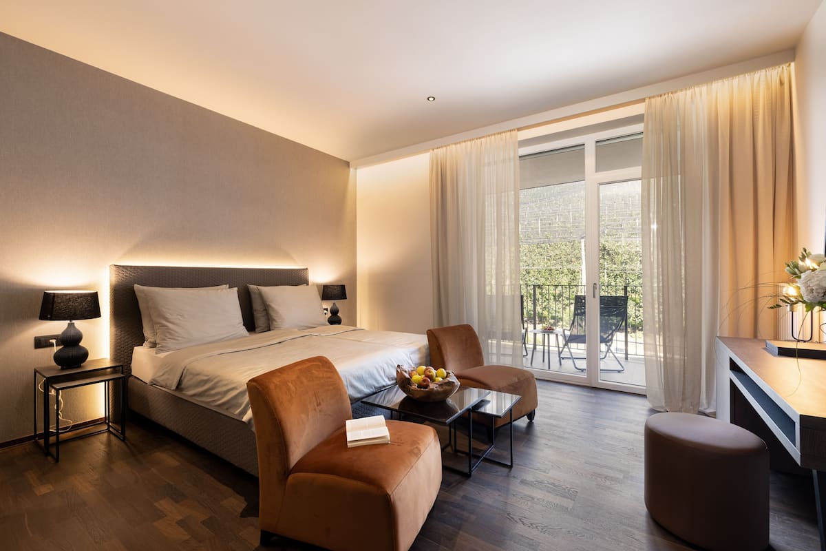 A modern hotel room with a large bed, a sofa, a small table with fruit, and a balcony with outdoor seating.