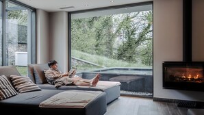 A modern living room with a large window, a fireplace, and a person reading a book. - Hotel Finkenhof (Schenna)