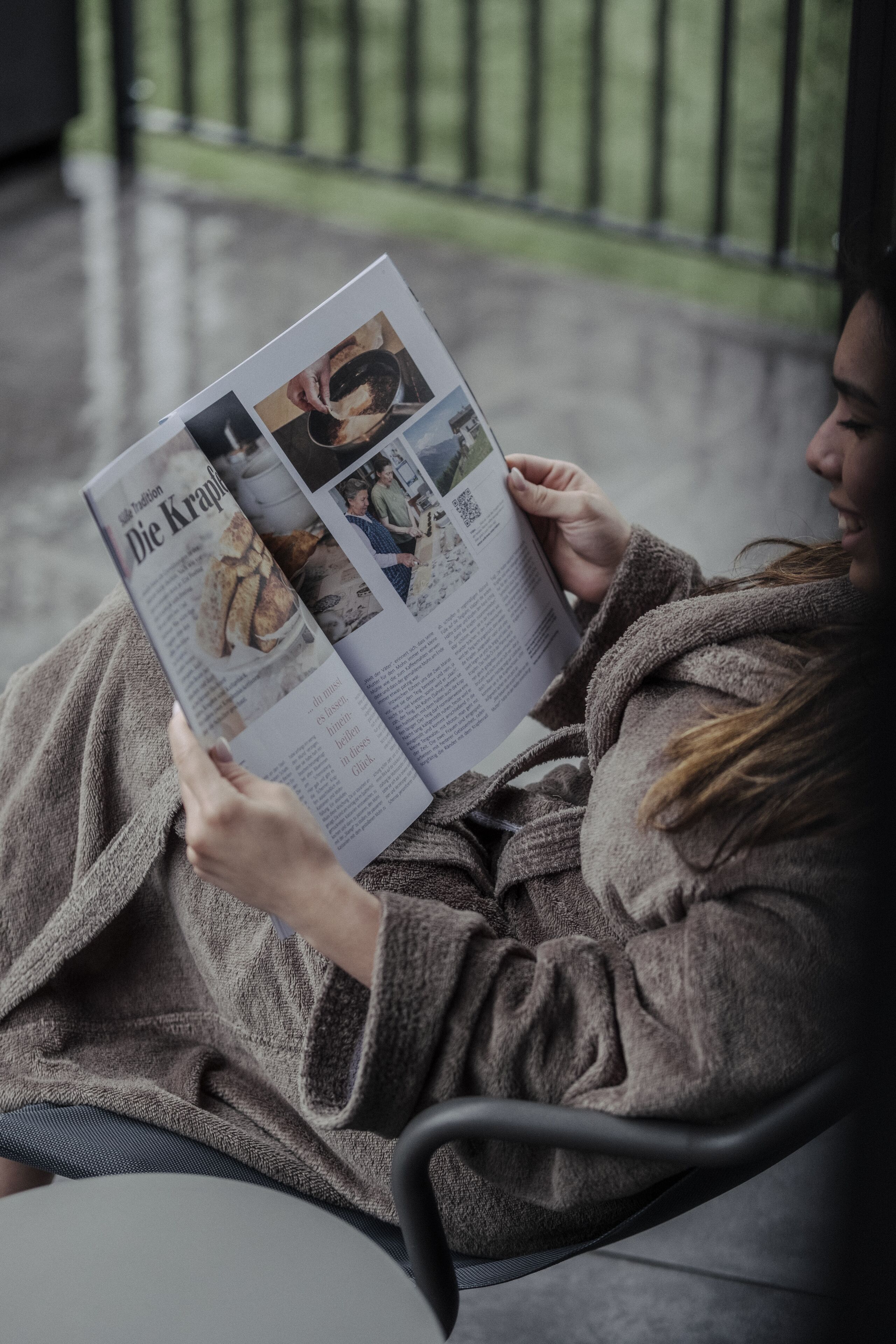 A person reading a magazine in a cozy chair with a view of a railing and greenery outside.