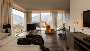 A hotel room with a bed, a TV, a desk with a lamp, a chair, and a view of mountains. - Hotel Finkenhof (Schenna)