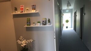 Un couloir aux murs blancs, une plante en pot posée sur une petite table, et des étagères remplies de brochures de voyage et de petites plantes.