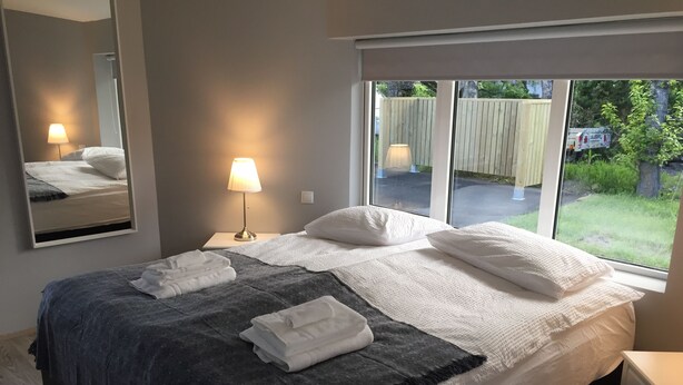 Un lit bien fait, avec du linge de lit blanc et gris, une lampe de chevet et une fenêtre donnant sur un jardin.