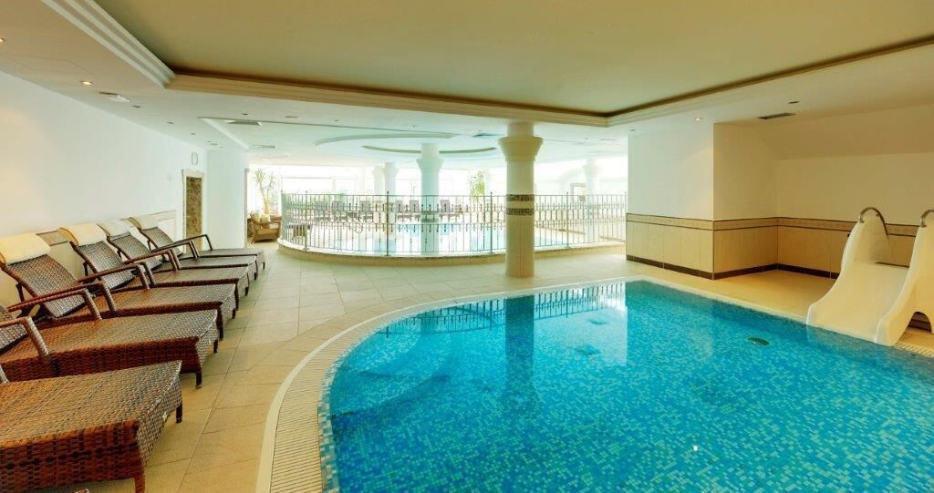 Una piscina interior con sillas de descanso y un tobogán, rodeada de suelo de baldosas y paredes blancas.