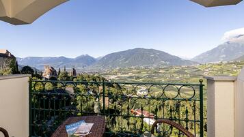 A balcony with wicker furniture and a magazine, overlooking a mountainous landscape with vineyards and a church.