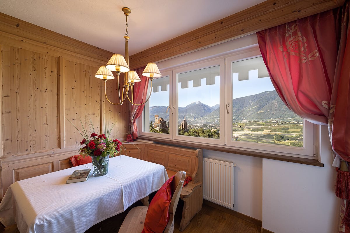 A room with a wooden paneled wall, a table with a white tablecloth, a vase of flowers, and a view of a mountainous landscape through a window with red curtains.