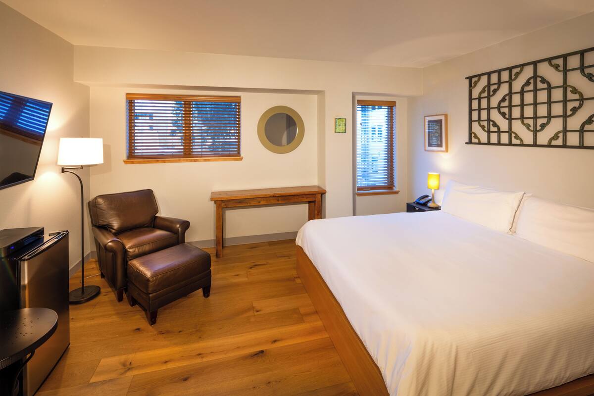 A hotel room with a bed, a leather chair, a wooden table, a desk, a lamp, a TV, and a wall-mounted artwork.
