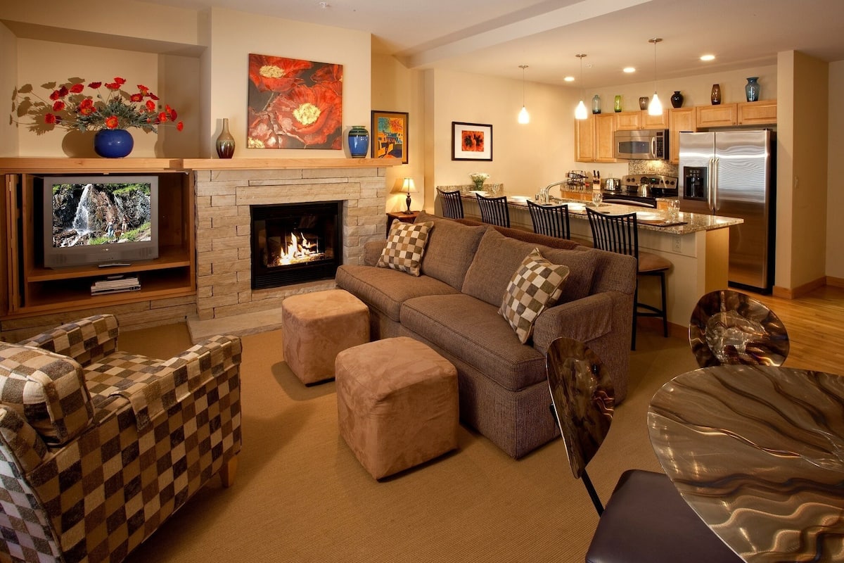 A living room with a fireplace, a television, a sofa, and a kitchen area with a refrigerator and microwave.