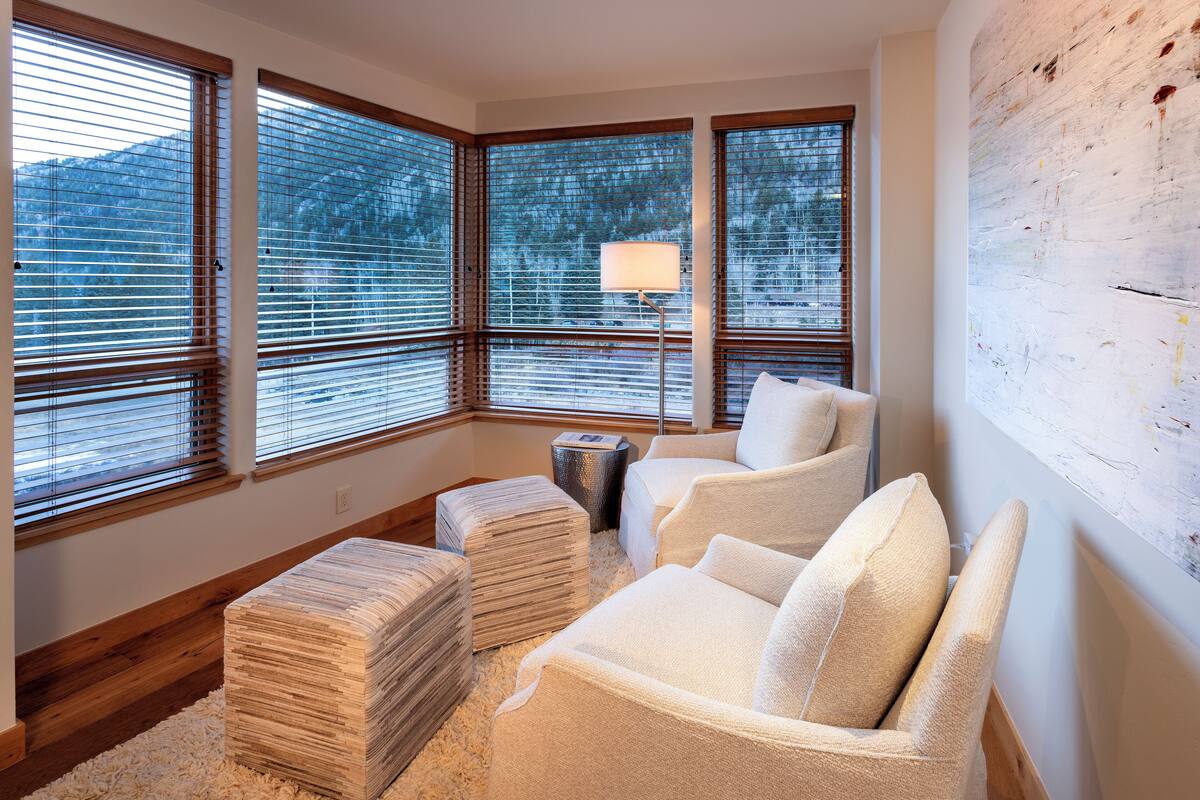 A corner room with large windows, a lamp, a white armchair, and a stack of books.