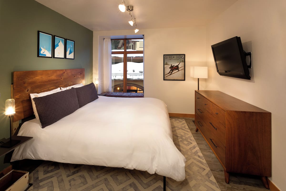 A neatly arranged bedroom with a large bed, a wooden dresser, a television, and framed artwork on the walls.