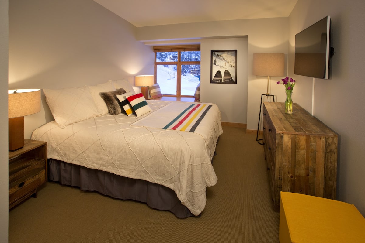 A bedroom with a bed, bedside lamps, a wooden dresser, a TV, and a window with a view of a snowy landscape.