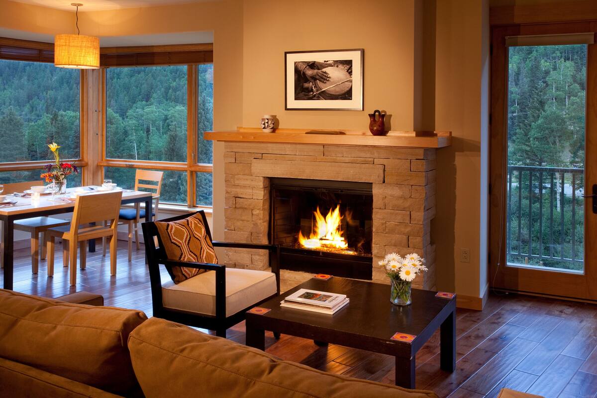 A cozy living room with a fireplace, wooden furniture, and large windows offering a forest view.