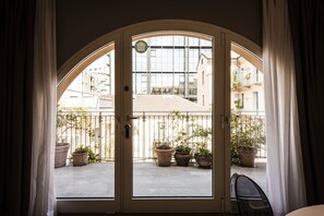 Uitzicht door een glazen deur met een metalen hekwerk, plantenpotten en gebouwen op de achtergrond.
