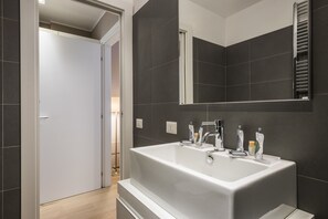 A modern bathroom with a large white bathtub, dark tiled walls, and a mirror above the sink. - MYSWEETPLACE - Corso Como Apartments (Milan)