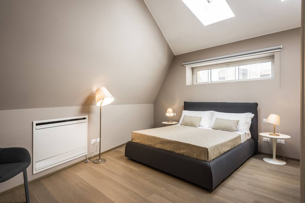 A modern bedroom with a large bed, two bedside lamps, a radiator, and a skylight.