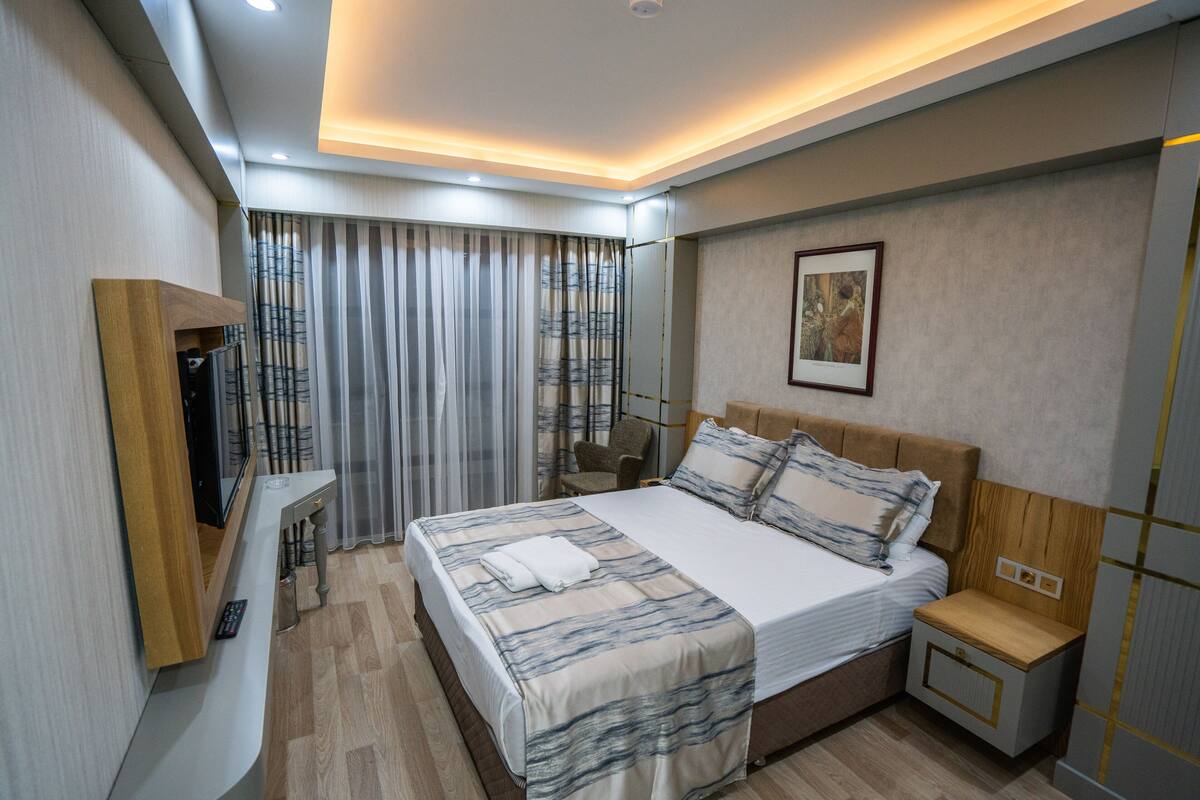 Una habitación de hotel moderna con una cama grande, mesitas de noche, un televisor y una ventana con cortinas.