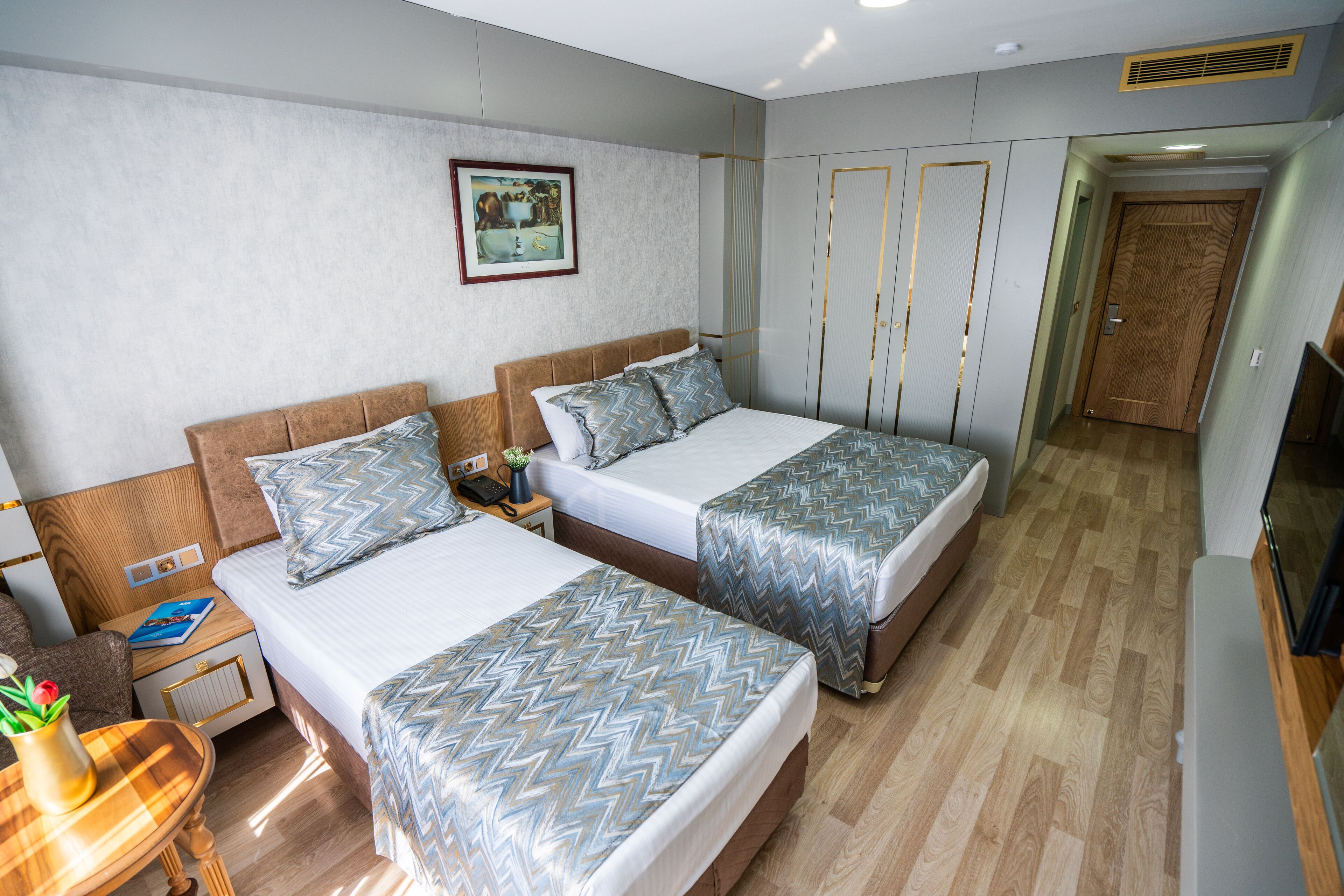 A hotel room with two beds, a wooden floor, a painting on the wall, and a door.