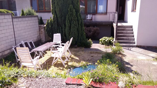 A backyard with a patio table and chairs, a small garden with plants and a birdbath, and a staircase leading to a second-floor balcony. - Large Apartment in the heart of Slovácko (Kunovice)