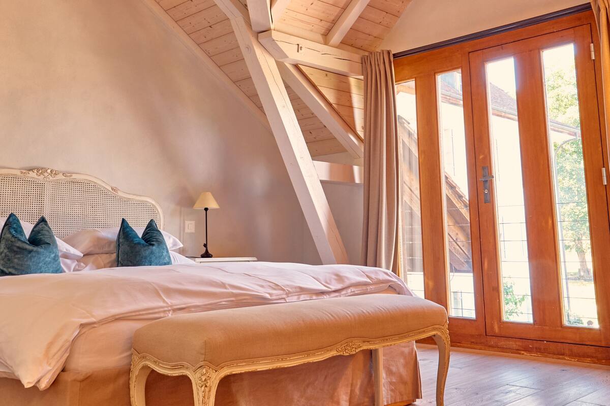 Een slaapkamer met een groot bed, houten balken, een raam en een deur.