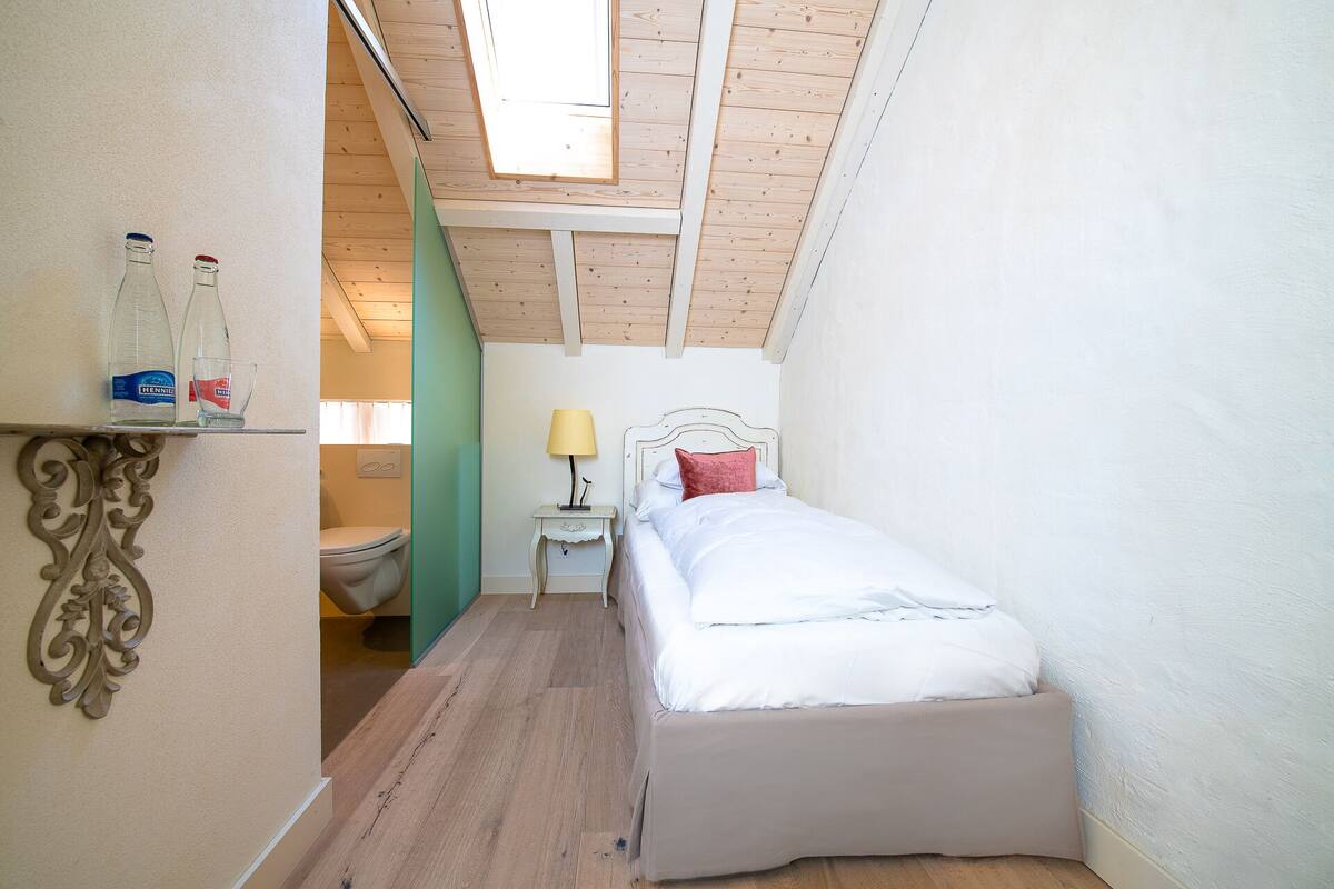 Een kleine kamer met een schuine kap, een bed met wit beddengoed, een nachtkastje met een lamp en een plank met flessen en glazen.