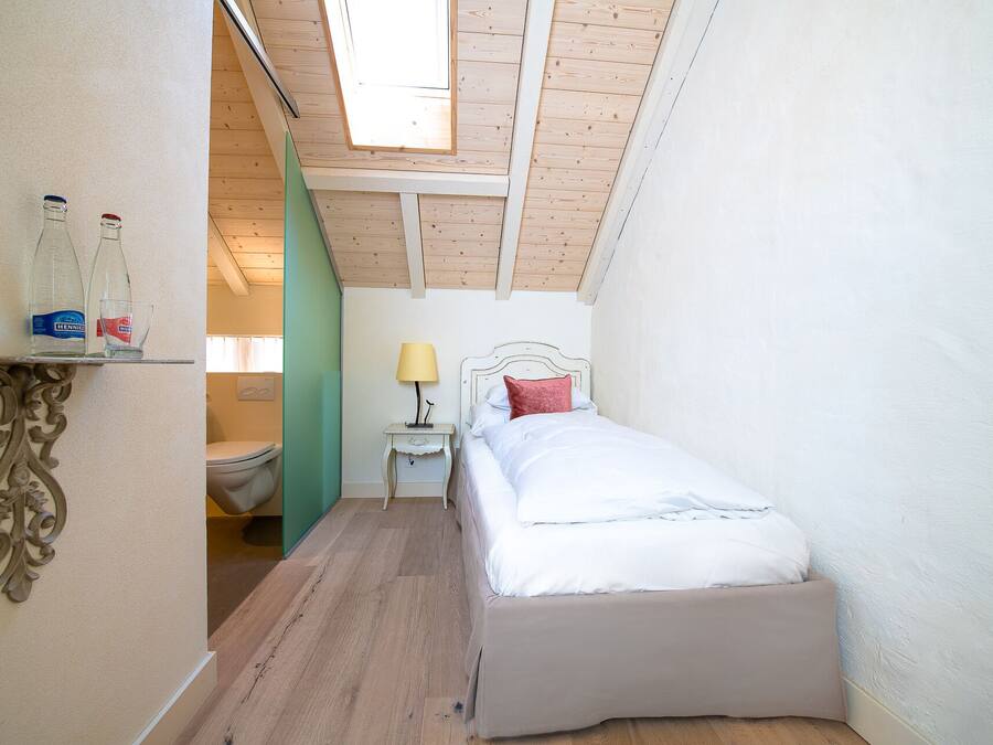 Una stanza piccola con soffitto spiovente, un letto con biancheria bianca, un comodino con una lampada e uno scaffale con bottiglie e bicchieri.