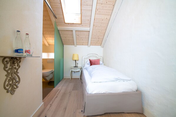 A narrow room with a sloped ceiling, a bed with white bedding, a bedside table with a lamp, and a shelf with bottles and glasses. - Chateau Salavaux (Vully-les-Lacs)