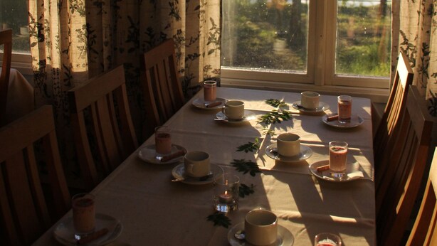 A dining table set for four with floral curtains and a view of trees and water outside. - Hotel Vuollerim (Vuollerim)