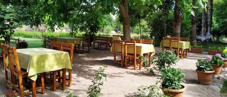 Un área de comedor al aire libre con mesas y sillas, rodeada de plantas en macetas y árboles.