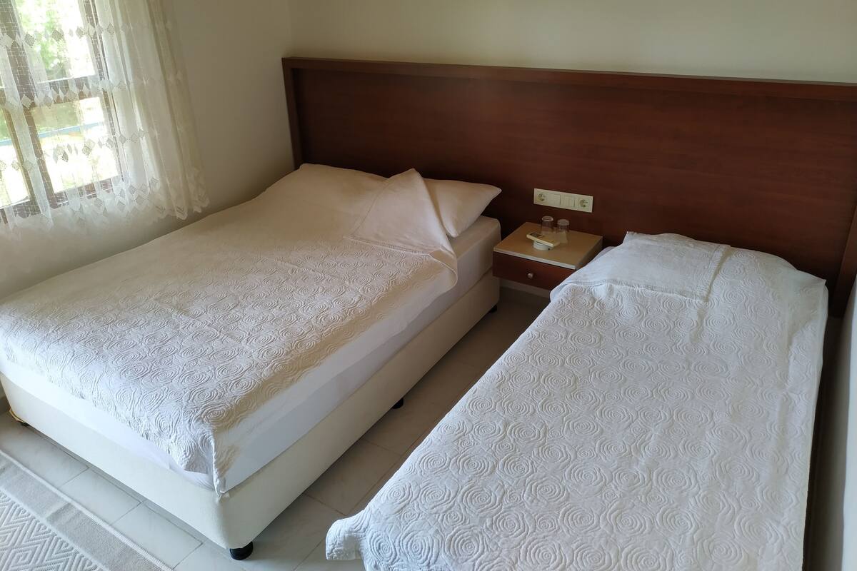 Ein Doppelbett mit Holz Kopfteil, ein Nachttisch mit Lampe und ein Fenster mit durchsichtigen Vorhängen.