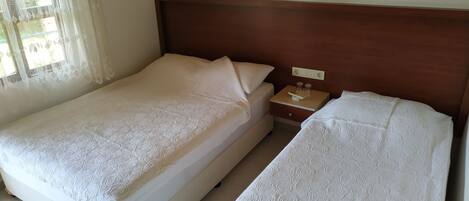 Una cama doble con cabecera de madera, una mesita de noche con lámpara y una ventana con cortinas de gasa.
