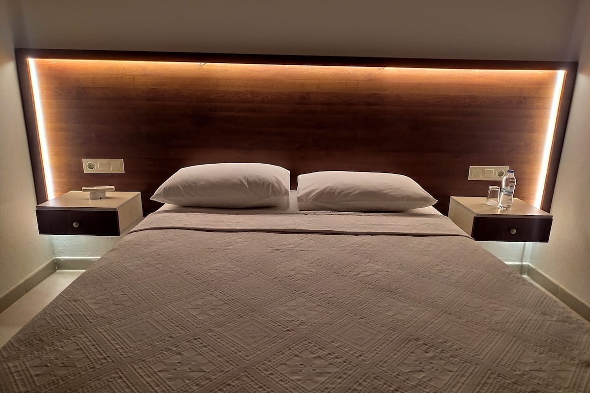 Ein ordentlich bezogenes Bett mit einem gemusterten Überwurf, zwei weißen Kissen und einem Holz-Kopfteil mit indirekter Beleuchtung. Es gibt zwei Nachttische, jeder mit einer kleinen Lampe und einer Wasserflasche.