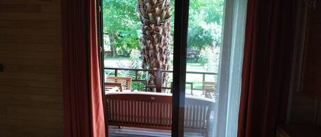 Habitación de hotel con cama, ventana con cortinas y vista a una terraza al aire libre con una palmera.