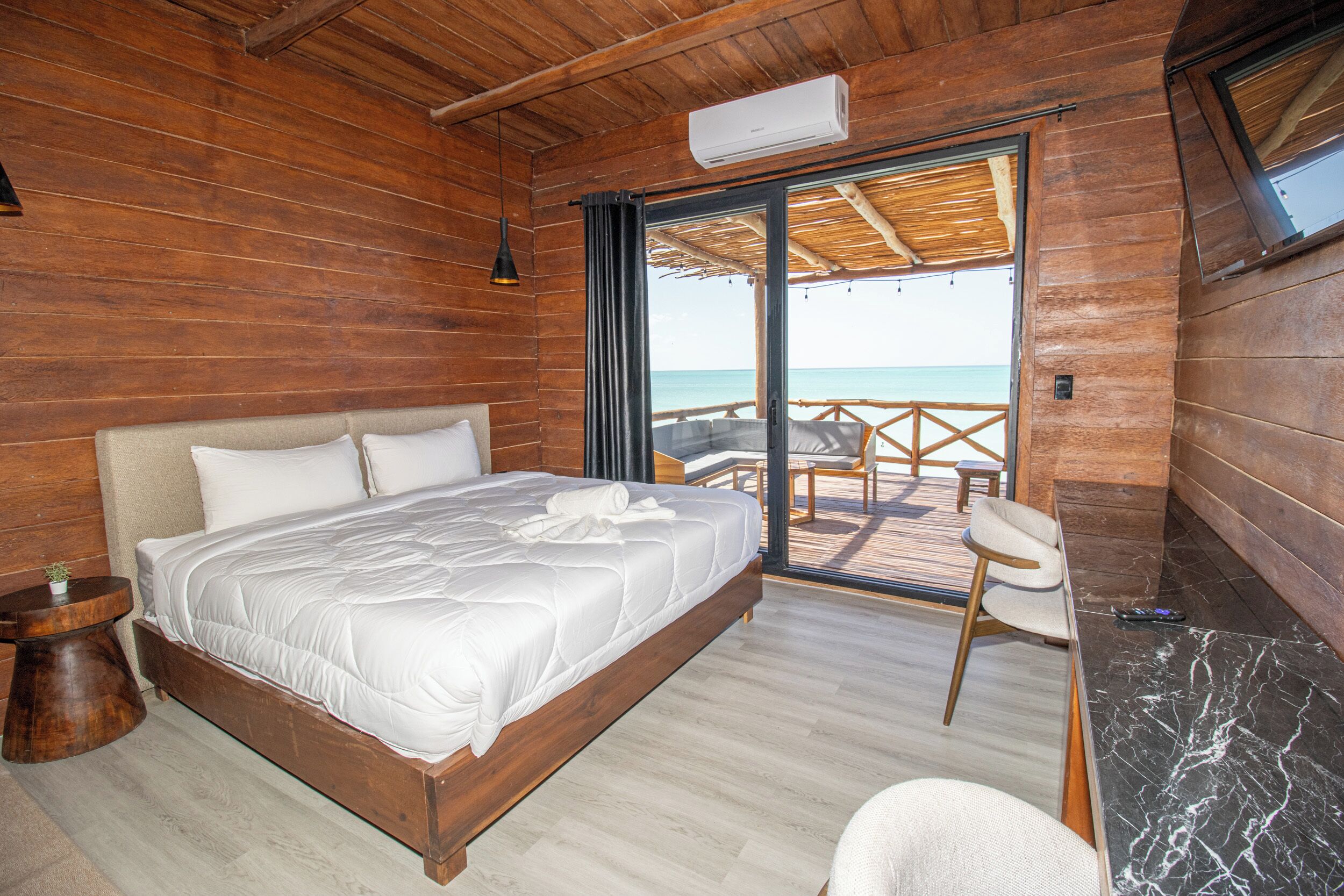 A wooden room with a bed, a small table, a chair, and a view of the sea through a sliding door.