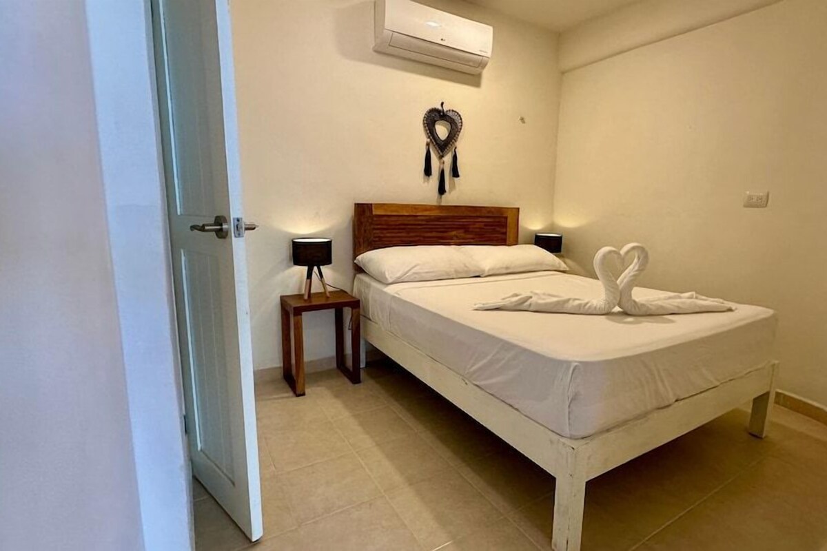 A bedroom with a bed, bedside table, wall-mounted air conditioner, and a heart-shaped decoration.