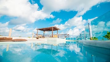 A clear blue pool with a wooden pergola and lounge chairs, surrounded by a modern building with large glass windows.