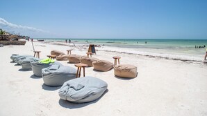Una playa con sillones de exterior y mesitas, cielo despejado y el océano al fondo.