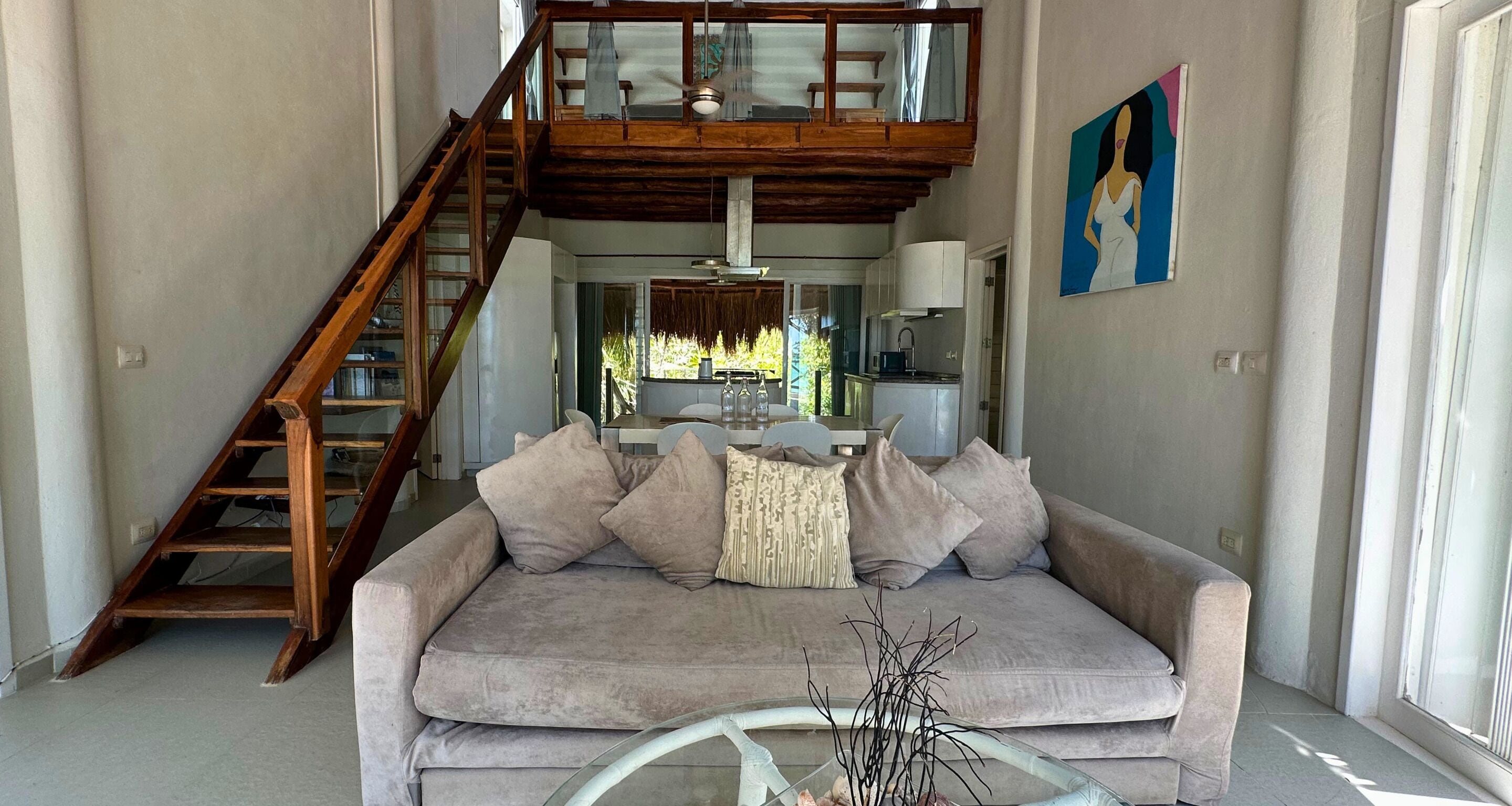A living room with a grey sofa, a glass coffee table, a wooden staircase, and a painting on the wall.