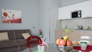 A modern living room with a grey sofa, a red chair, and a glass table with fruit and a glass of water. - Alludo B&B (Agrigento)