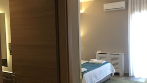 A hotel room with a wooden door, a bed with a blue blanket, a white air conditioning unit, and a green chair. - Alludo B&B (Agrigento)