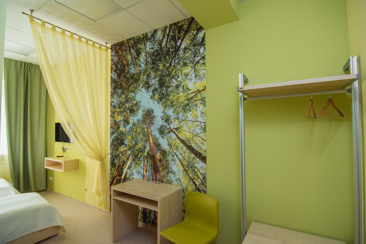 Une chambre d’hôtel équipée d’un lit, d’un bureau, d’une chaise, d’un miroir, d’une étagère, d’une armoire et d’une grande fresque représentant une forêt sur le mur.
