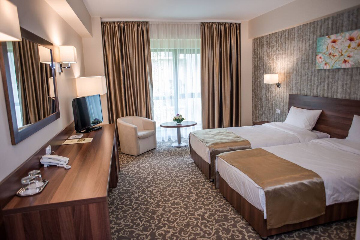 A hotel room with a wooden desk, two beds, a chair, a small table with a plant, and a large window with curtains.