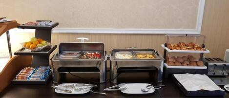Una mesa de buffet con diversos alimentos, como frutas, pasteles y platos calientes, dispuestos con utensilios de servicio.